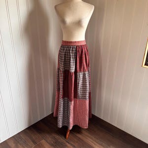 May include: A long, floor-length skirt featuring a patchwork design of various red, white, and grey plaid and gingham fabric panels. The skirt has a fitted waistband with a red and white checkered pattern and a full, flowing silhouette.