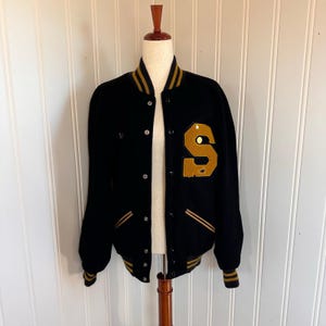 May include: Black varsity jacket with a large gold "S" patch on the front. The jacket has gold and black striped trim on the collar, cuffs, and waist. The jacket is buttoned up and displayed on a mannequin.