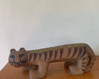 Op de afbeelding: Een keramiek tijgerbeeldje met bruine, groene en zwarte strepen. De tijger zit in een hurkende positie met zijn kop naar de zijkant gedraaid.