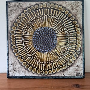 May include: A square ceramic tile with a raised, textured design. The centre of the tile features a circle of small blue and white dots surrounded by a ring of raised, yellow, teardrop-shaped forms. The tile is framed with a dark grey border. The tile is signed "Lisa L. Denmark".