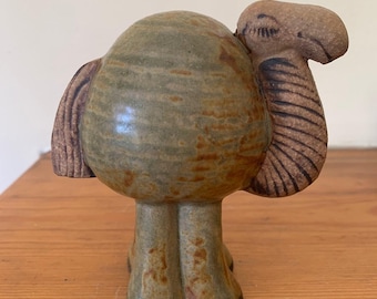 May include: A ceramic camel figurine with a green and brown glaze. The camel has a rounded body and short legs.