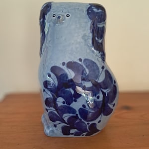 Może przedstawiać: Ceramiczna figurka psa w odcieniach niebieskiego. Pies ma jasnoniebieski korpus z ciemnoniebieskimi wzorami i akcentami kwiatowymi. Figurka stoi na drewnianej powierzchni.