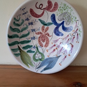 May include: A white ceramic bowl with a floral design. The bowl features a variety of flowers in shades of pink, blue, green, and orange. The flowers are painted in a stylised manner, with some appearing to be more abstract than others.