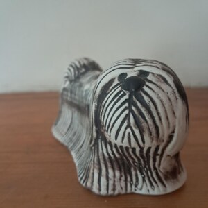 Może przedstawiać: Ceramiczna figurka psa, z białą podstawą i czarnymi paskami i detalami. Pies ma czarny nos i oczy, a sierść jest teksturowana. Figurka stoi na drewnianej powierzchni.