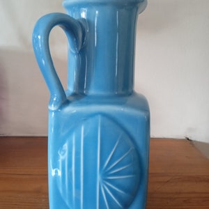 Puede incluir: Una jarra de cerámica azul claro con base cuadrada y asa. La jarra tiene un diseño decorativo en la parte delantera, con un sol estilizado.