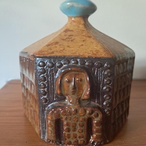Puede incluir: Una caja hexagonal de cerámica con una figura humana estilizada en la parte delantera. La caja tiene un esmalte marrón y beige con una tapa esmaltada en azul.