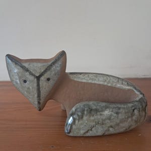 Può includere: Una statuetta di volpe in ceramica con un design stilizzato. La volpe è marrone e grigia con accenti neri. È seduta su una superficie di legno.