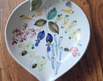 Può includere: Un piatto da portata in ceramica bianca a forma di foglia con un manico. Il piatto è decorato con un motivo floreale in tonalità di blu, verde, giallo, rosa e marrone.