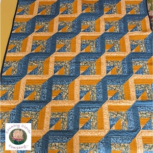 May include: A quilt with a geometric pattern in shades of blue and orange. The quilt is made up of squares and triangles, and the pattern creates a 3D effect. The quilt is finished with a label that reads "Country Clover Company."