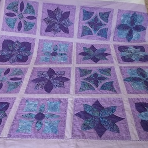 Può includere: Una trapunta viola e blu con un motivo floreale. La trapunta è composta da 16 quadrati, ognuno con un design diverso. La trapunta è su uno sfondo bianco.