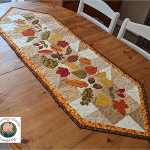 Peut inclure: Un chemin de table matelassé avec un motif de feuilles d'automne dans des tons de brun, orange et jaune. Le chemin de table a une bordure brune, orange et jaune. Le texte "Country Clover Company" est dans le coin inférieur gauche de l'image.