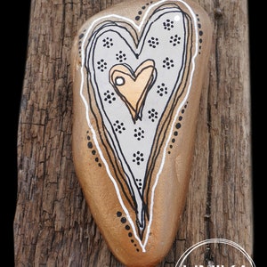 Pode incluir: Uma pedra em forma de coração pintada com um coração dourado dentro de um coração cinza com pontos pretos e brancos. A pedra está montada em um pedaço de madeira desgastada com o texto "UNIKA DESIGN" na parte inferior.