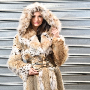 May include: A long, brown and white fur coat with a large hood. The coat has a belt with a buckle and is made of a soft, luxurious material.