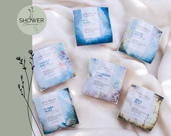 Puede incluir: Seis vaporizadores de ducha individuales, cada uno en un paquete cuadrado con un diseño y nombre únicos. Los paquetes presentan fondos de estilo acuarela en tonos de azul, morado y dorado, con etiquetas de texto. La imagen incluye el texto "SHOWER Steamers".