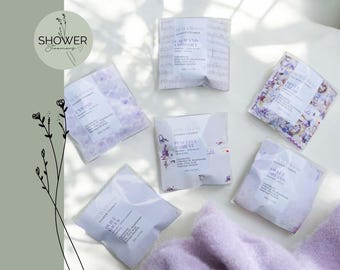 Puede incluir: Vista cenital de seis vaporizadores de ducha en envases transparentes. Cada paquete lleva una etiqueta blanca con texto y detalles florales. La paleta de colores es suave, con tonos lavanda y blanco, creando un ambiente relajante.