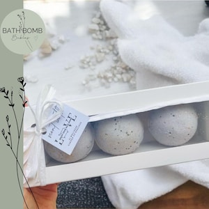 Puede incluir: Una caja blanca que contiene tres bombas de baño moteadas. La caja está atada con una cinta blanca y una etiqueta que dice "scent of love". Las bombas de baño son de color gris claro con motas oscuras. El fondo incluye una toalla blanca y elementos decorativos.