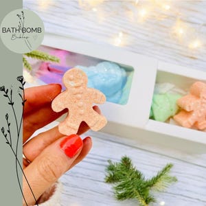 May include: A hand holds a gingerbread man bath bomb. The bath bomb is light brown. A white box contains other bath bombs in blue, green, and orange. The text "BATH BOMB Bubbles" is in a circle in the upper left. Festive bath products.
