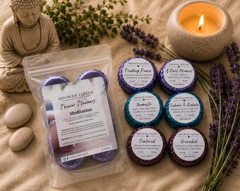 Puede incluir: Una disposición de vaporizadores de ducha, una vela encendida y ramitas de lavanda. Los vaporizadores de ducha son de varios colores y están etiquetados con frases como "Finding Peace" y "Namaste". También hay una estatua de Buda y piedras lisas.