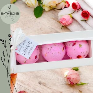 May include: A white box containing three pink bath bombs with rose petals. The box is tied with a white ribbon and a tag that reads "scent with LOVE". The image includes roses and a towel.