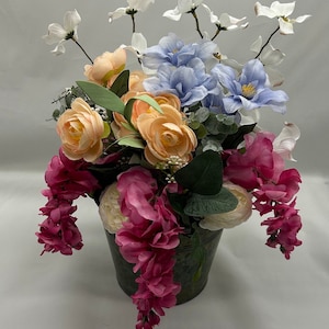 May include: A floral arrangement in a metal bucket with pink, peach, white, and blue flowers. The flowers are arranged in a cascading style.