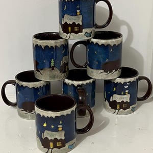 May include: Set of six blue and brown ceramic mugs with a winter scene design. Each mug features a snowy house, a tree, and stars against a dark blue background. The mugs have brown handles and a brown rim.