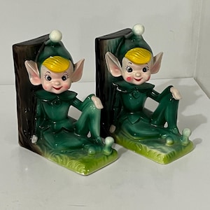 May include: A pair of vintage ceramic elf bookends. Each elf is dressed in a green outfit with a matching hat and yellow hair. The bookends are designed to look like a tree trunk and grass.