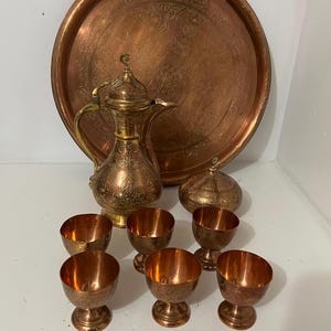 May include: A copper-toned serving set featuring a large round tray with etched floral designs, a decorative pitcher, a lidded bowl, and six small footed cups. The set has a vintage aesthetic and is likely used for serving beverages or food.