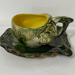 May include: A ceramic teacup and saucer set. The cup and saucer are green with a textured, shell-like design. The interior of the cup is yellow. The saucer has a wavy edge. A unique and decorative addition to any tea set collection.