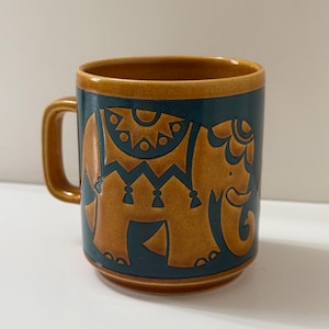 May include: A ceramic mug with a teal background and a brown elephant design. The mug has a brown handle and rim. The elephant is decorated with intricate patterns. The mug is a standard size for coffee or tea.