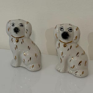 Puede incluir: Dos figuras de perro de cerámica blanca con ojos y narices negros, collares dorados y manchas doradas. Los perros están sentados, con las patas delanteras en el suelo. Decorativo.