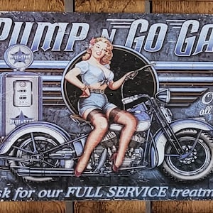 Könnte beinhalten: Ein Vintage-Schild aus Metall mit einem Pin-up-Girl auf einem Motorrad, einer Zapfsäule und dem Text "Pump N Go Gas" und "Open all Night". Das Schild sagt auch "ask for our FULL SERVICE treatment".