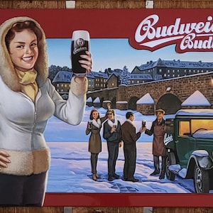 Könnte beinhalten: Ein Vintage-Metallschild, das für Budweiser Budvar Bier wirbt. Das Schild zeigt eine Frau in einem weißen Mantel, die ein Glas Bier hält, und einen grünen Lastwagen im Hintergrund. Das Schild ist vor einer verschneiten Stadtkulisse platziert.
