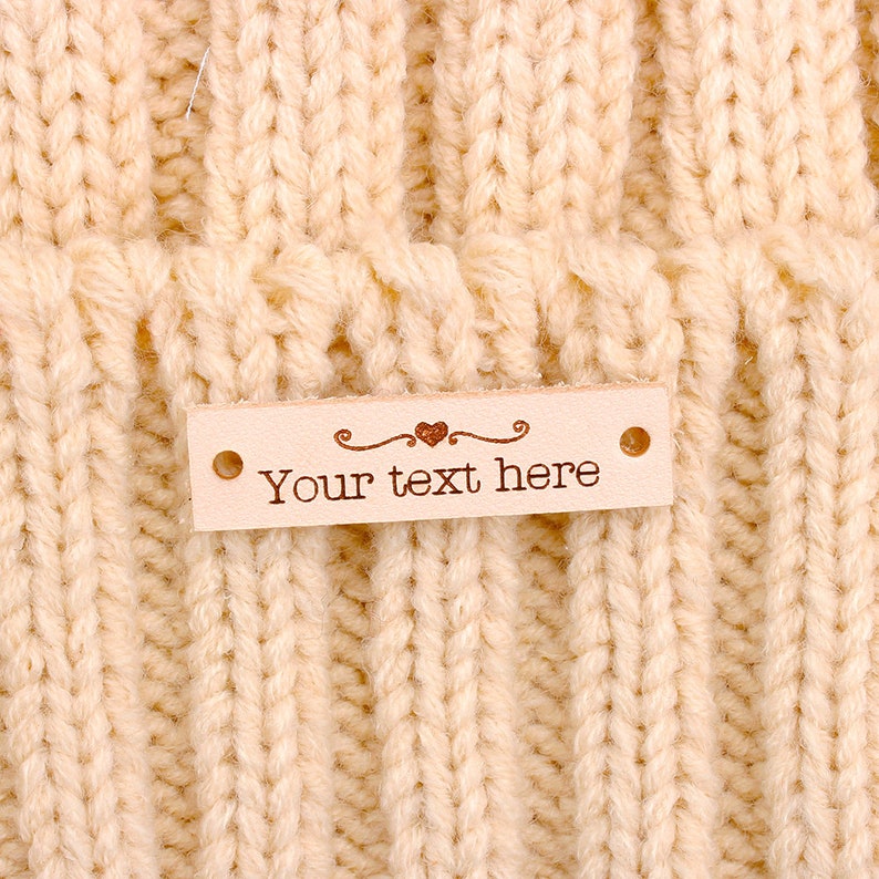 May include: A light brown leather tag with a heart design and the text "Your text here". The tag has two holes on the sides and is attached to a knitted fabric.