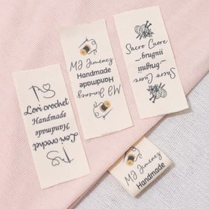 May include: A collection of off-white fabric labels with black text and illustrations, arranged on a pink surface. The labels display text including "Lori crochet Handmade" and "MJ Jimenez Handmade". Some labels also feature a heart symbol and a thread spool.