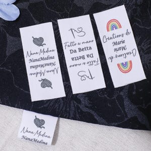 May include: Multiple white fabric labels with text and illustrations. One label reads "Nana Medina" with a yarn heart graphic. Another says "Fatto a mano Da Betta". A third label displays "Creations de Marie" with a rainbow.