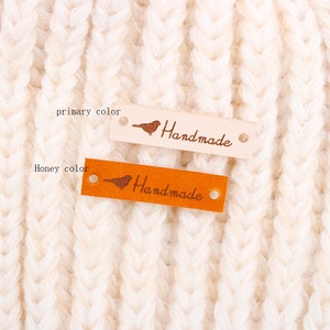 May include: Two leather labels with a small bird design and the word "Handmade" in a script font. The label on the left is a natural colour and the label on the right is a honey colour.