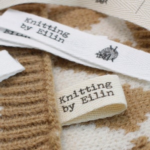 May include: Close-up of a white cotton fabric label with black text that reads "Knitting by Eillin". The label is attached to a brown and white knitted fabric.