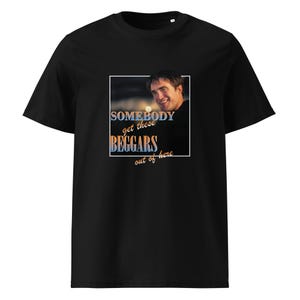 Può includere: Una t-shirt nera a maniche corte con una stampa grafica quadrata di una persona sorridente. Il testo SOMEBODY get these BEGGARS out of here è scritto con un gradiente blu e arancione, incorniciato da un sottile bordo bianco sul petto.