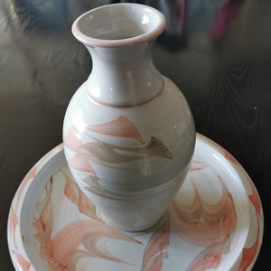 Könnte beinhalten: Eine weiße Keramikvase mit rundem Körper und schmalem Hals, die auf einem passenden runden Teller steht. Beide Teile sind mit abstrakten, fließenden Designs in Pfirsich- und Brauntönen verziert. Das Vasen- und Teller-Set ist ein dekoratives Wohnaccessoire.