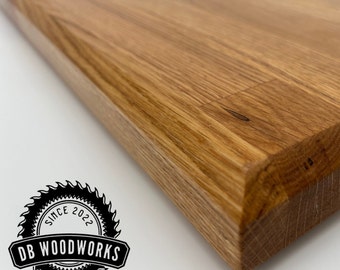 May include: A close-up of a natural wood cutting board with a beveled edge. The board is made of light brown wood with a grain pattern. The board is a rectangular shape and has a smooth surface.
