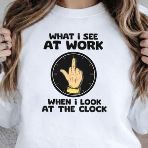 May include: White t-shirt with the text "WHAT I SEE AT WORK" above a clock graphic. The clock shows a hand giving the middle finger. Below the clock is the text "WHEN I LOOK AT THE CLOCK".
