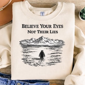 May include: A cream-coloured sweatshirt featuring the text "BELIEVE YOUR EYES NOT THEIR LIES" above a black and white illustration of a penguin standing before mountains. The penguin is facing away.