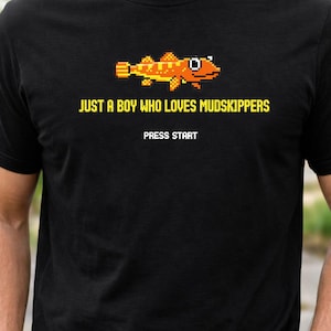 May include: Black t-shirt featuring a pixelated orange and yellow mudskipper design. The text reads "JUST A BOY WHO LOVES MUDSKIPPERS" above "PRESS START" in yellow. A fun, casual tee for those who love mudskippers.