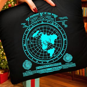 May include: Black square pillow with a teal map of the world design. The pillow features the text "NEW STANDARD MAP OF THE WORLD" and other text. The pillow is being held in front of a Christmas tree and bookshelf.