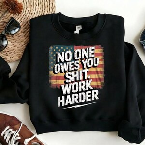 Puede incluir: Sudadera negra con el texto blanco "NO ONE OWES YOU SHIT, WORK HARDER" sobre un diseño de bandera americana desgastada. La sudadera se muestra con gafas de sol, zapatos marrones y vaqueros.