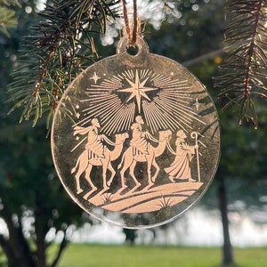 Puede incluir: Un adorno navideño de vidrio transparente con una silueta blanca de los tres reyes magos montando camellos y una estrella encima.