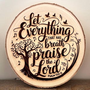 Puede incluir: Una rodaja redonda de madera con un diseño quemado de un árbol y las palabras "Que todo lo que tiene aliento alabe al Señor Salmo 150:6".