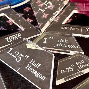 Może przedstawiać: Przezroczyste akrylowe szablony do pikowania z wymiarami w calach, w tym "1.5 Half Hexagon" i "1.25 Half Hexagon". Widoczne są również pomarańczowe nożyczki i niebieski długopis, a także różowa i czarna tkanina.