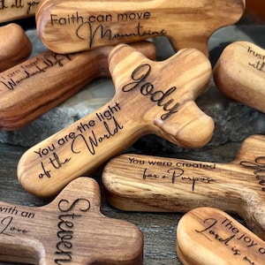May include: A collection of wooden crosses with various engraved inspirational phrases and names. The crosses are made of light and dark brown wood, with visible grain patterns. Some phrases include "Trust in the Lord," and "Faith can move mountains."