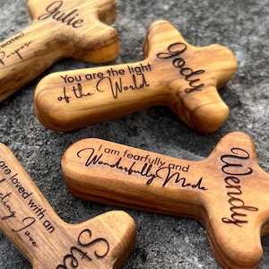 May include: Four wooden cross-shaped keepsakes with engraved names and inspirational quotes. The crosses are made of light brown wood and rest on a gray stone surface. The names Julie, Jody, Wendy, and Steven are visible.